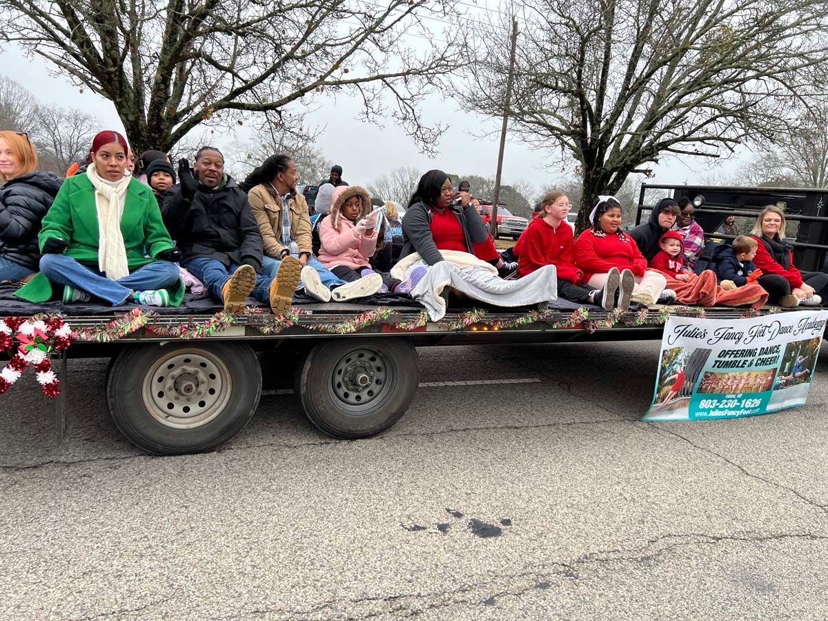 People seated on a trailer float in a parade, wearing colorful clothes and Christmas decorations.