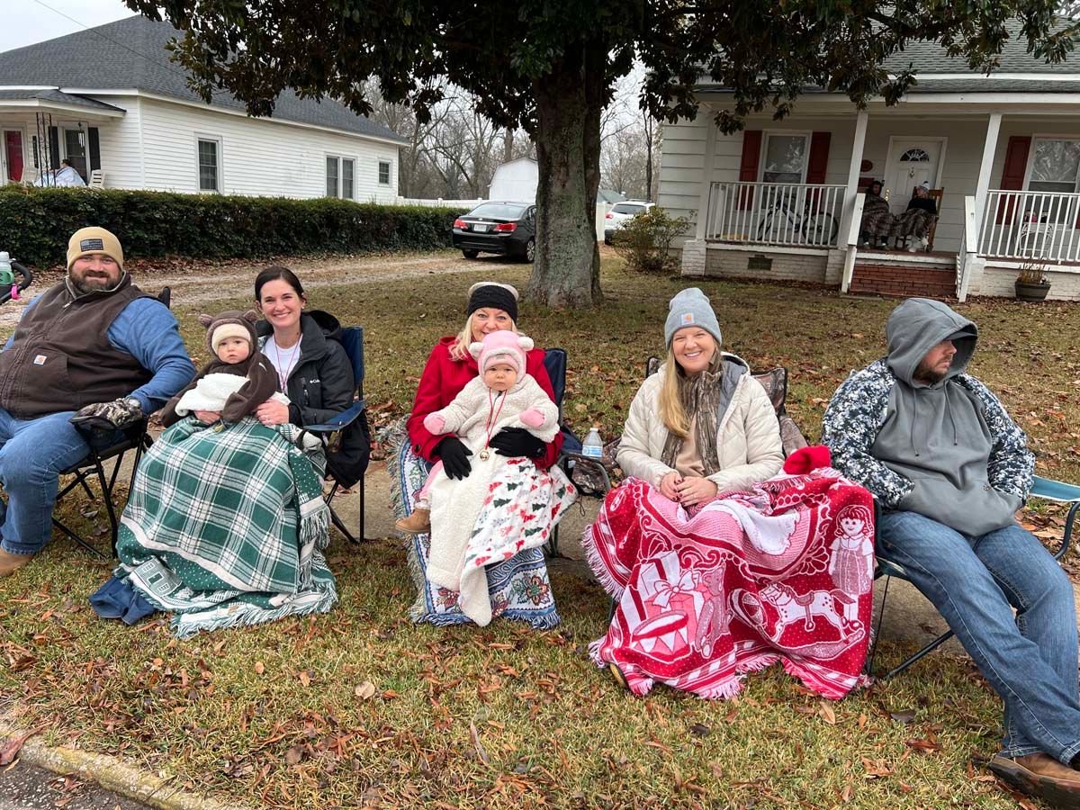 People seated outdoors on blankets and chairs under a tree, wrapped in winter coats and hoodies.