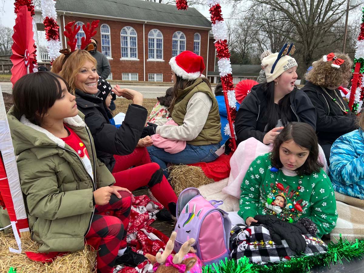 People in festive holiday attire ride on a red parade float in front of a brick building.