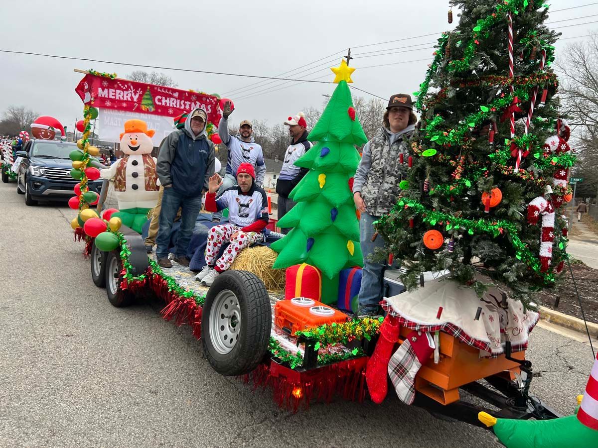 Decorated holiday parade float with Christmas trees, ornaments, lights, and people riding along the street