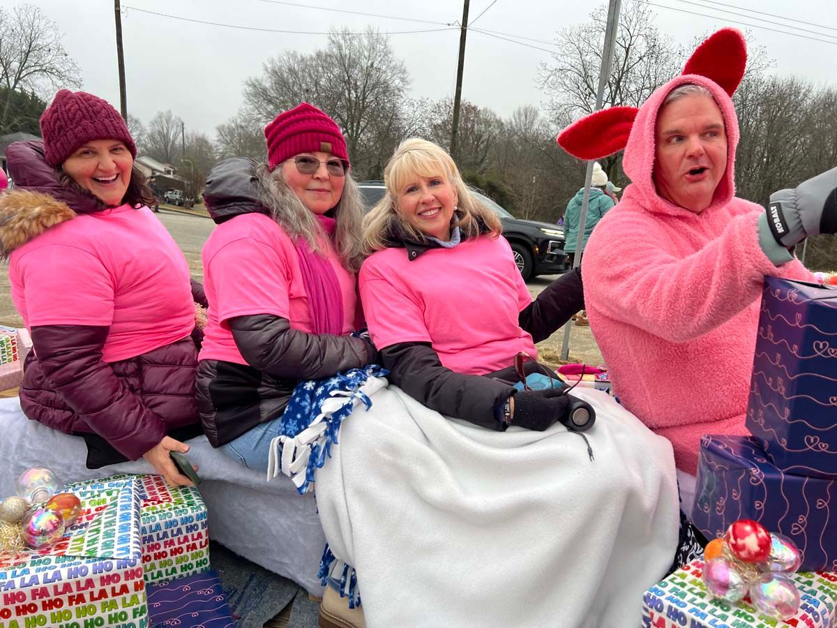 Four people in pink outfits ride on a decorated parade float, smiling in a snowy outdoor scene