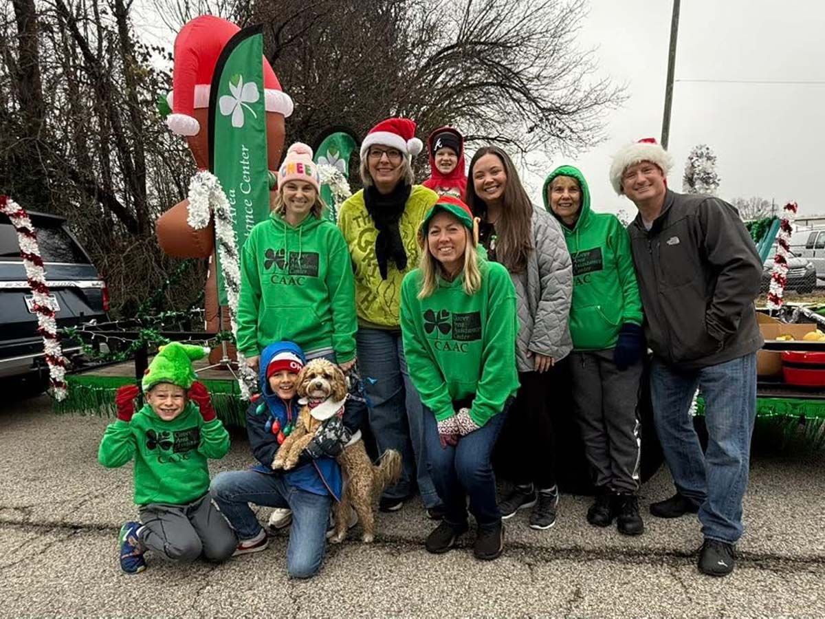 Group in green holiday shirts posing outdoors with a giant candy cane decoration and decorated vehicles.