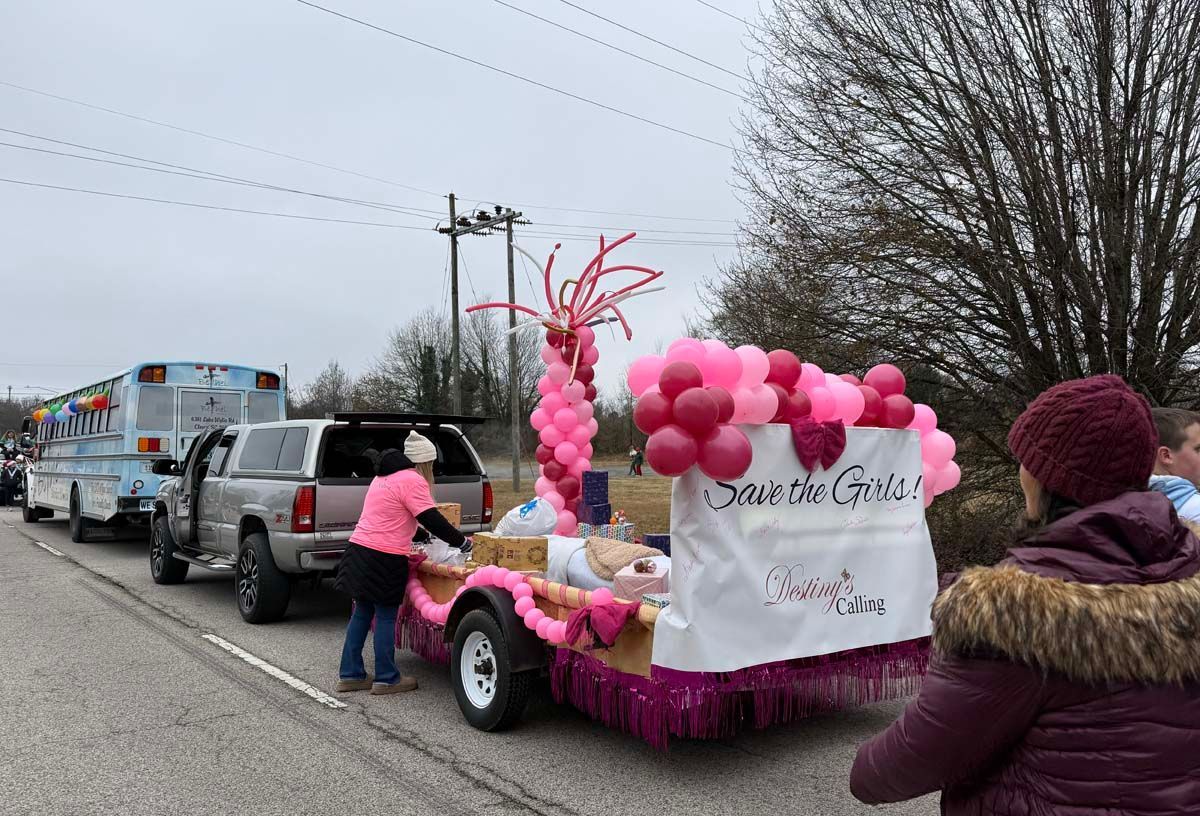 Pink-decorated parade float with banner “Sweet n’ Crafty” on a roadside, pulled by a truck, with people walking alongside
