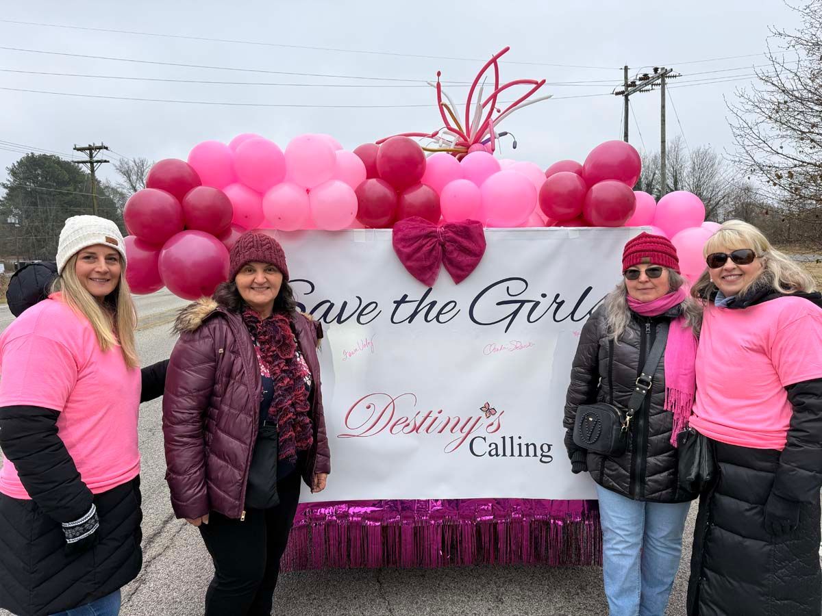 Four women pose beside pink flowers and a sign reading “Save the Girls” and “Destiny Calling” on a cloudy day