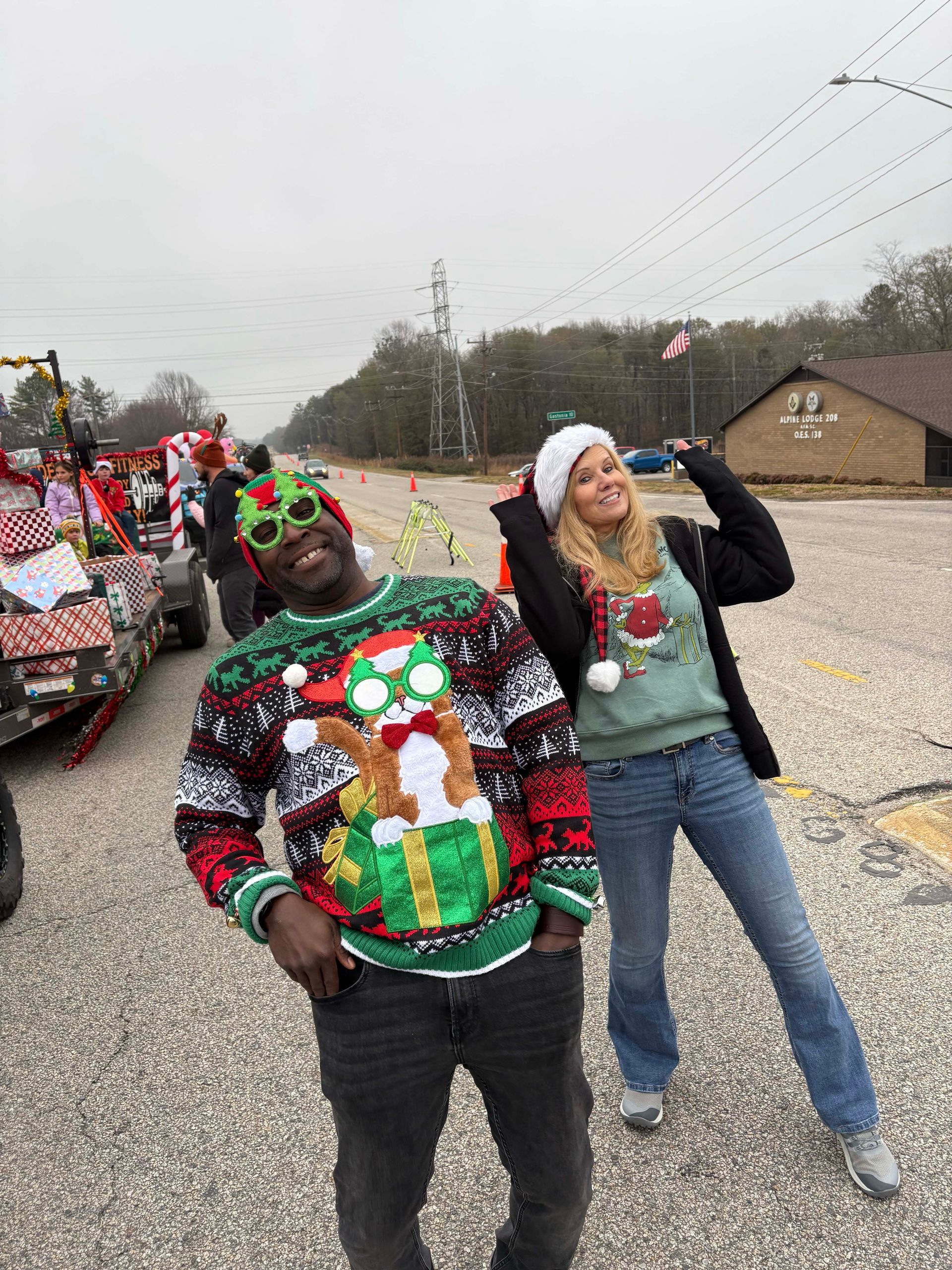 Two people in festive holiday sweaters posing outdoors during a parade, one waving and smiling.