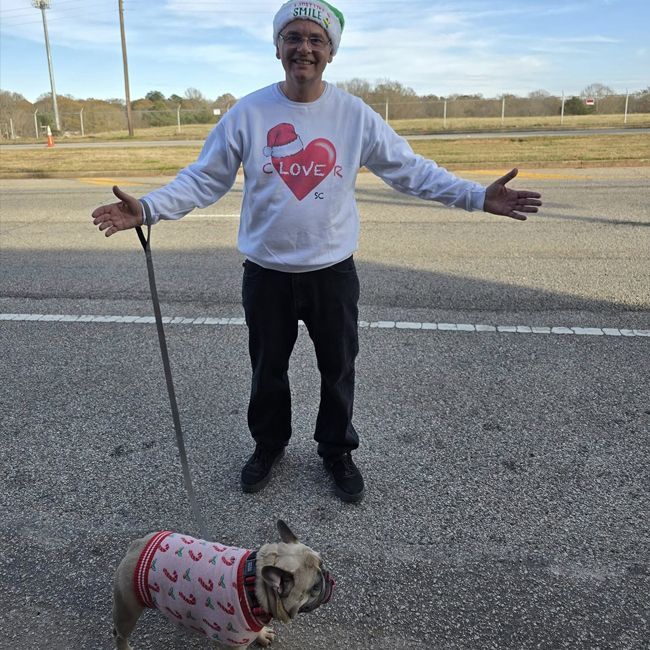 Person standing on a road with arms outstretched beside a small dog in a sweater.