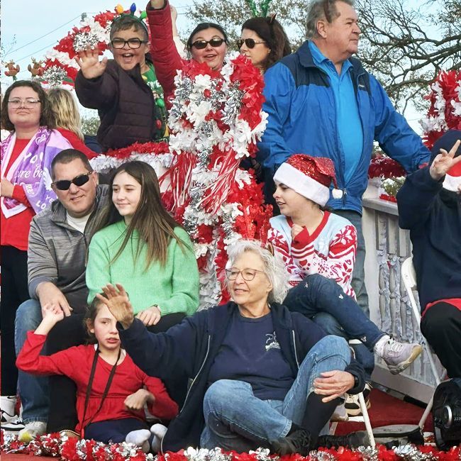 Group of people posing outdoors on a holiday float, decorated with red and white garlands and a Santa hat.