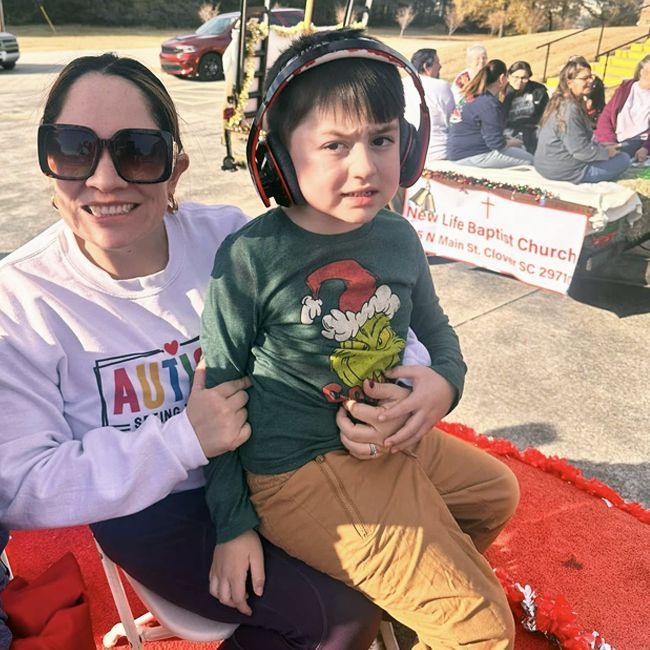 Woman holding child seated on red carpet at outdoor holiday event, both wearing headphones and smiling.