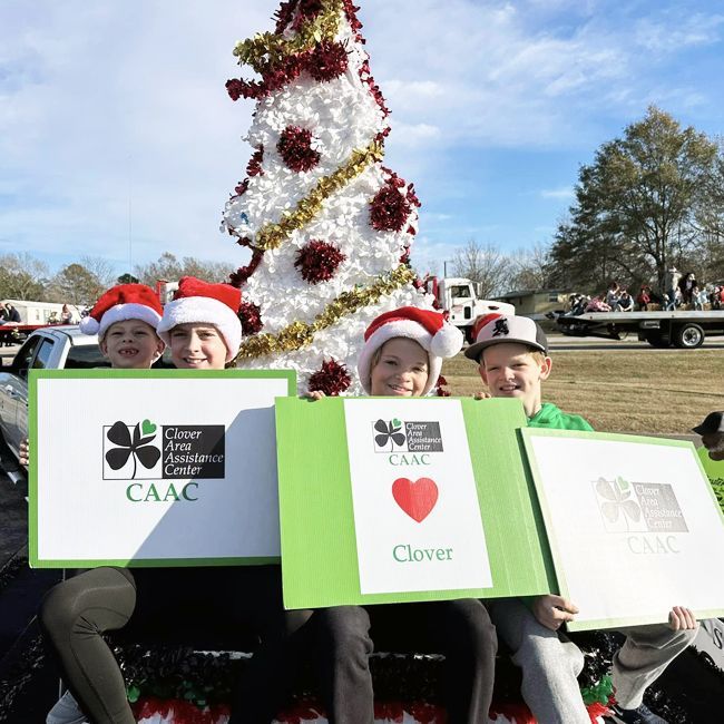 Children in Santa hats holding holiday signs beside a decorated tree at an outdoor event