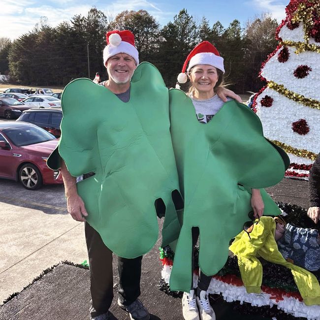 Two people in Santa hats wearing green tree costumes pose by a decorated outdoor holiday display.