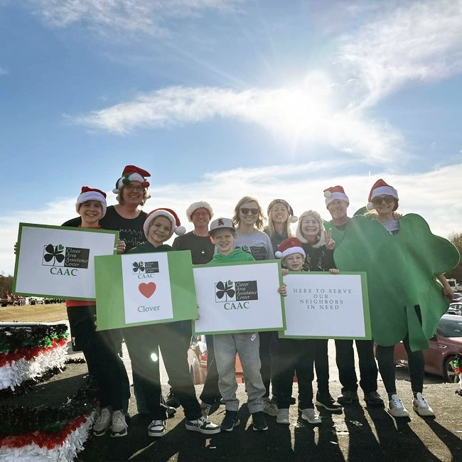Group of people in Santa hats holding signs outdoors in bright sunlight, with one person in a green costume.