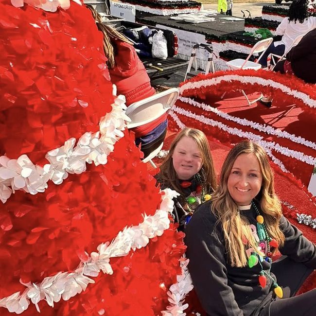 Two women smiling in a bright red holiday parade float with white trim and festive decorations