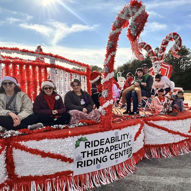 People on a red-and-white parade float for Rideability Therapeutic Riding Center, decorated with flowers and streamers.