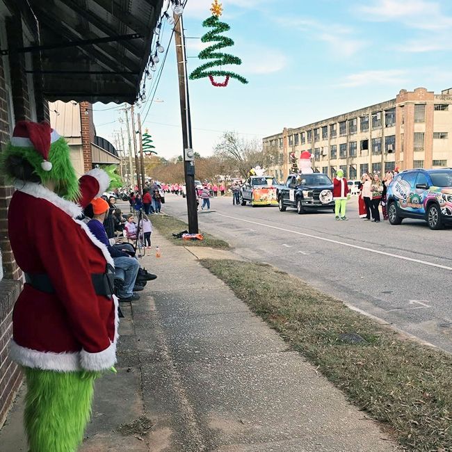 Holiday parade with a Grinch in red Santa coat watching cars decorated for Christmas on a city street