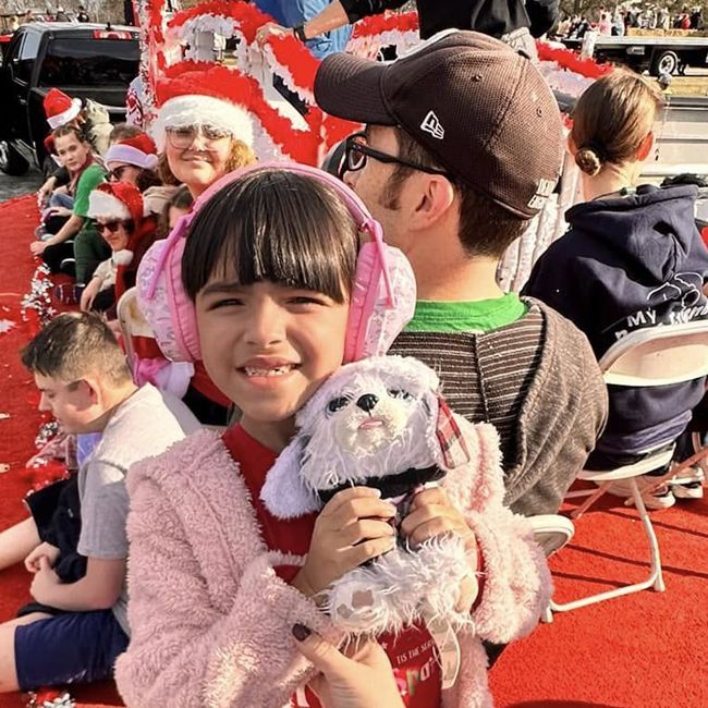 Smiling child in pink coat holding a plush dog at a festive outdoor parade with Santa in the background