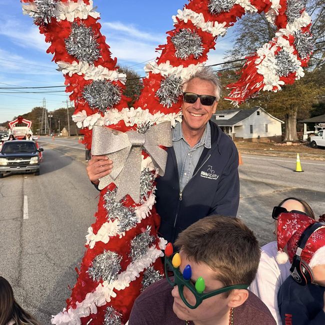Man smiling beside a red-and-silver holiday parade float on a street, with a child in the foreground.