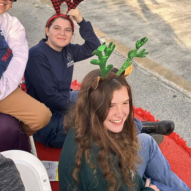 Two people in festive antler headbands sit outdoors, smiling on a red-and-gray paved surface.