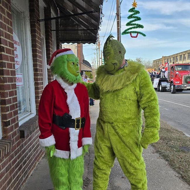 Two people in Grinch costumes standing on a sidewalk, one dressed as Santa, with a Christmas tree sign overhead.