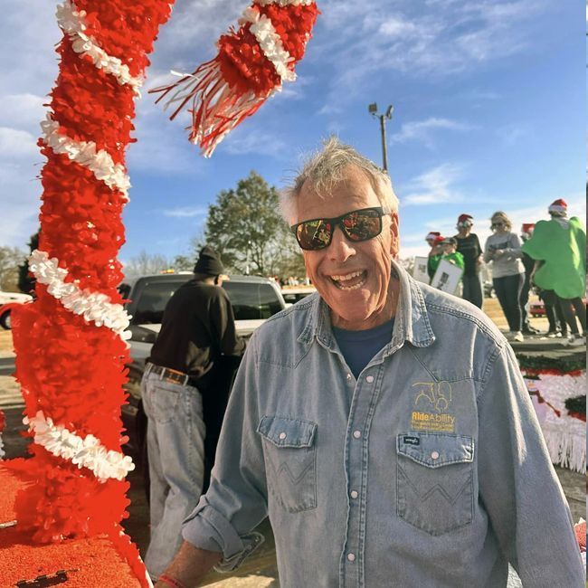 Smiling man in sunglasses at an outdoor event beside a red-and-white decorated post under a blue sky