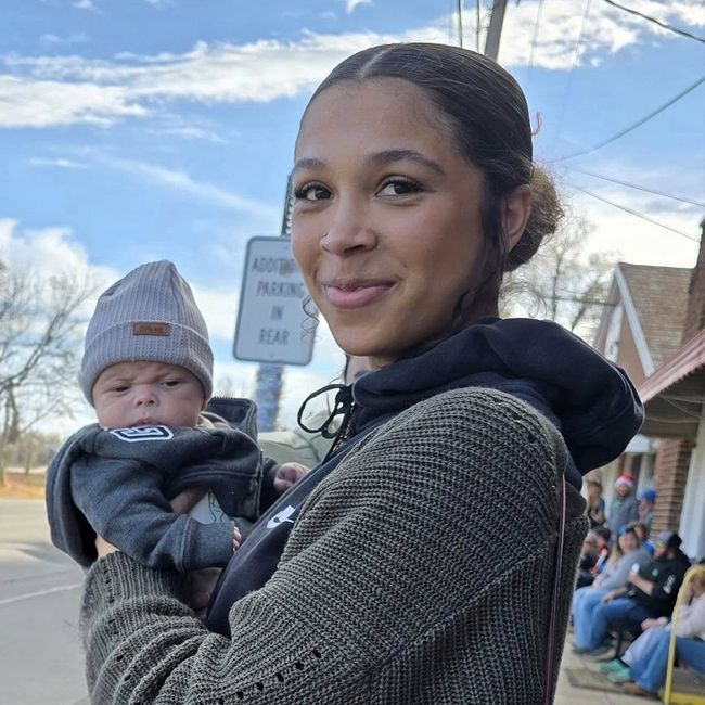 Person holding a baby outdoors on a sidewalk, both wearing dark clothing and looking at the camera.