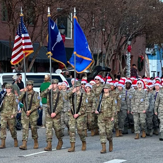 Military parade with uniformed soldiers marching behind U.S. and state flags, some wearing Santa hats.