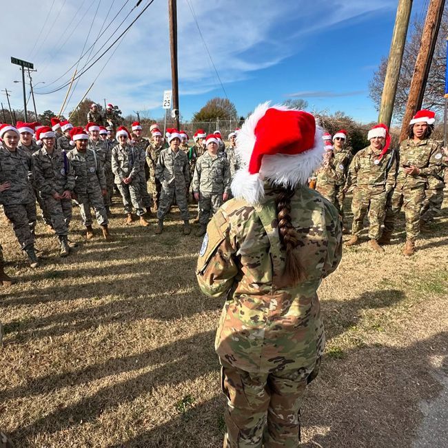 Soldiers in camouflage wearing Santa hats stand in formation outdoors on a sunny day.