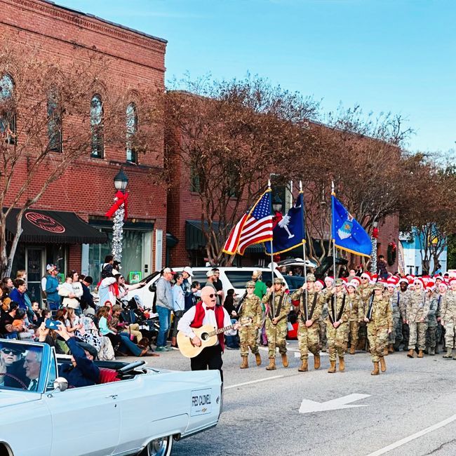 Parade marching past a brick storefront with flags, soldiers, and a white convertible carrying a guitarist