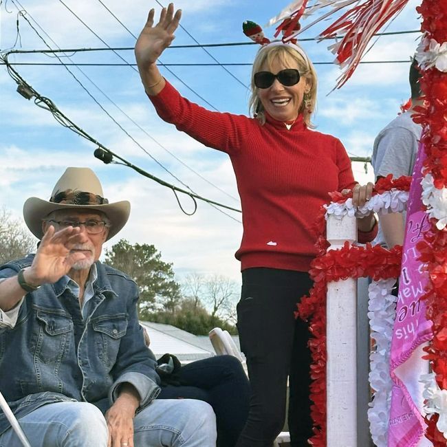 Two people in a decorated parade float waving, one in a cowboy hat and one in a red sweater and sunglasses.
