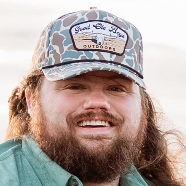 Smiling person in a camouflage cap and teal shirt with long hair and beard outdoors