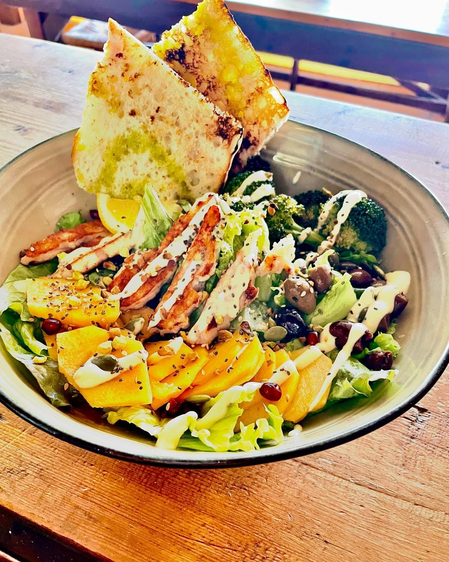 Ciotola colorata di cereali con verdure a foglia verde, verdure arrostite, pollo a fette e pane piatto su un tavolo di legno.