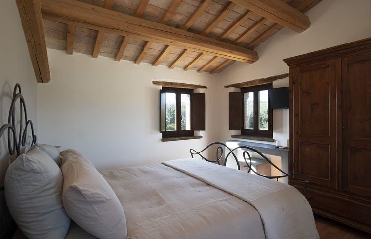Camera da letto rustica con soffitto con travi a vista, due finestre, letto e armadio.