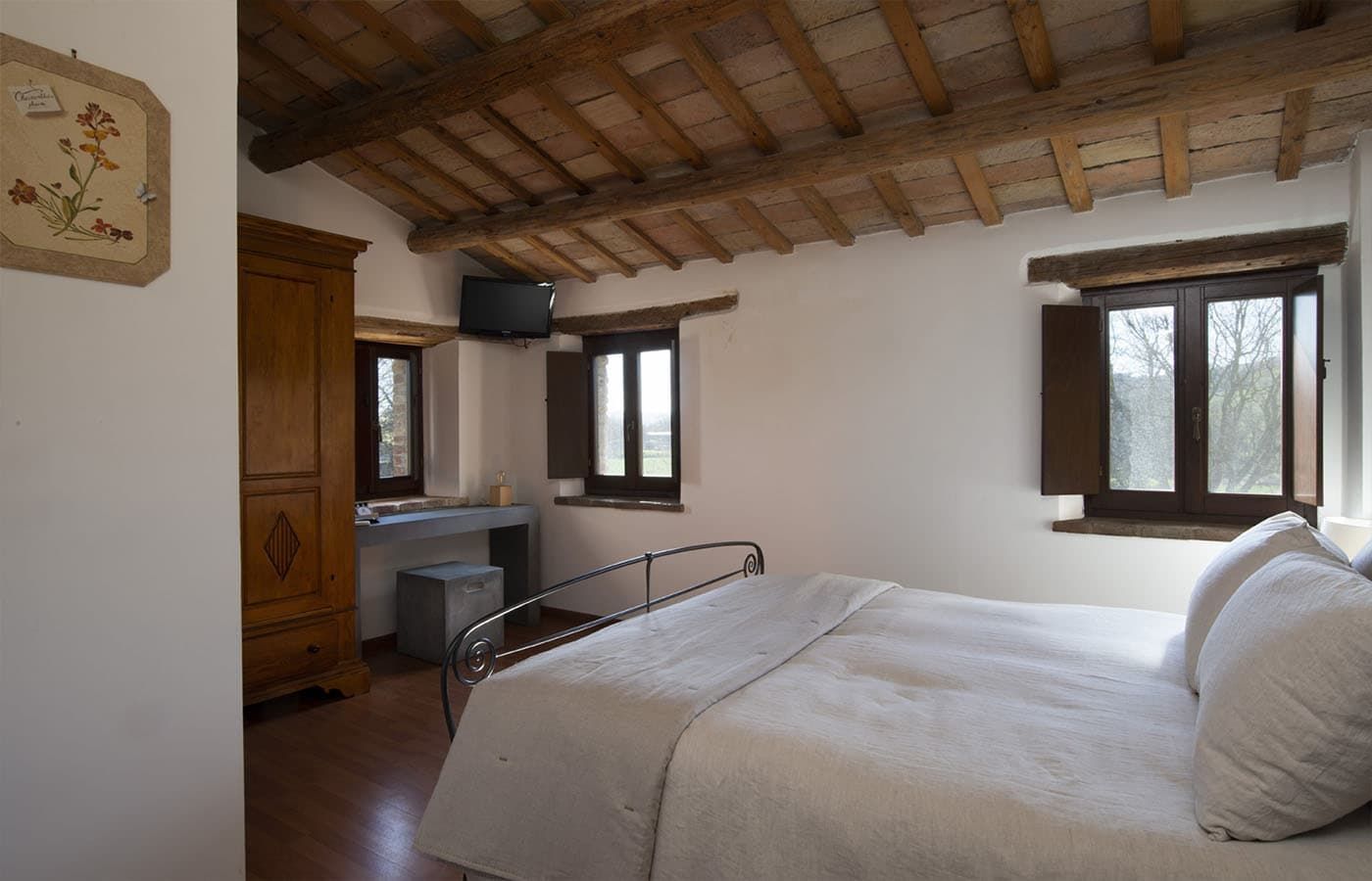 Camera da letto rustica con letto bianco, travi in ​​legno a vista e due finestre che si affacciano sugli alberi.