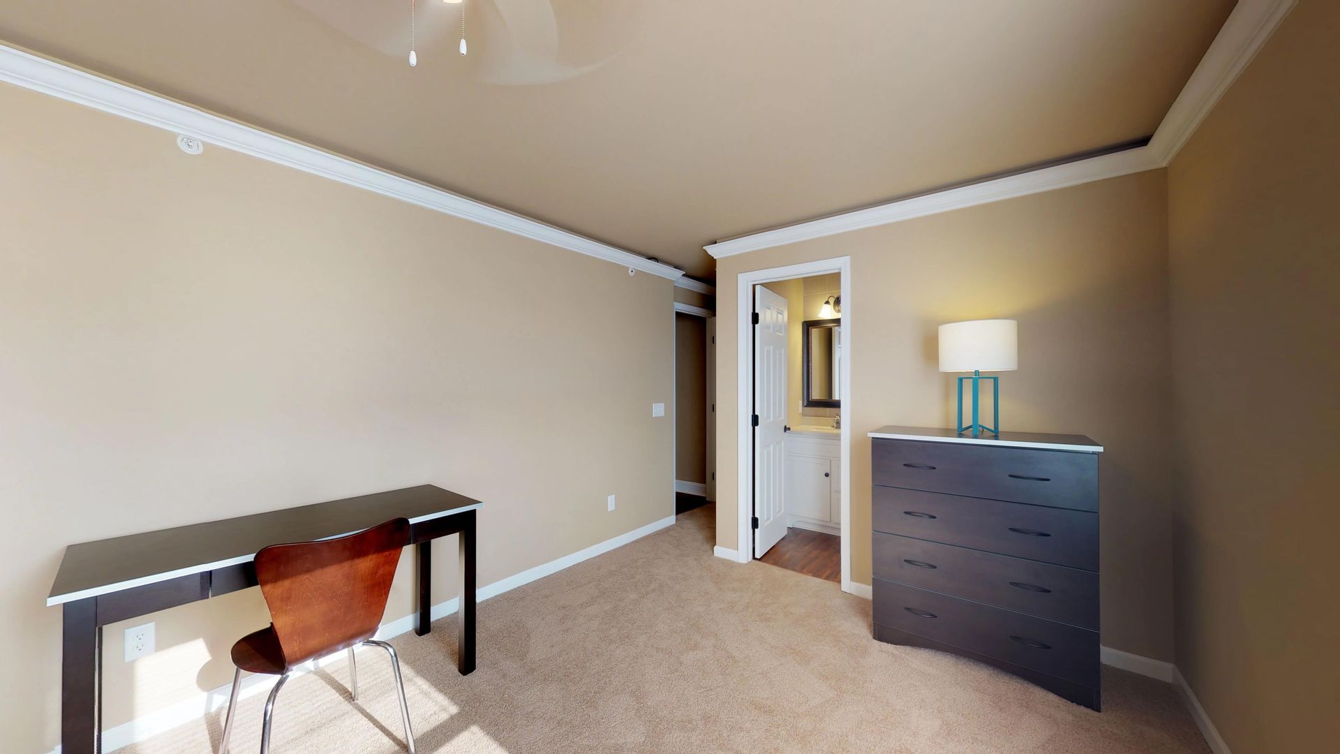 A bedroom with a desk , chair , dresser and lamp.
