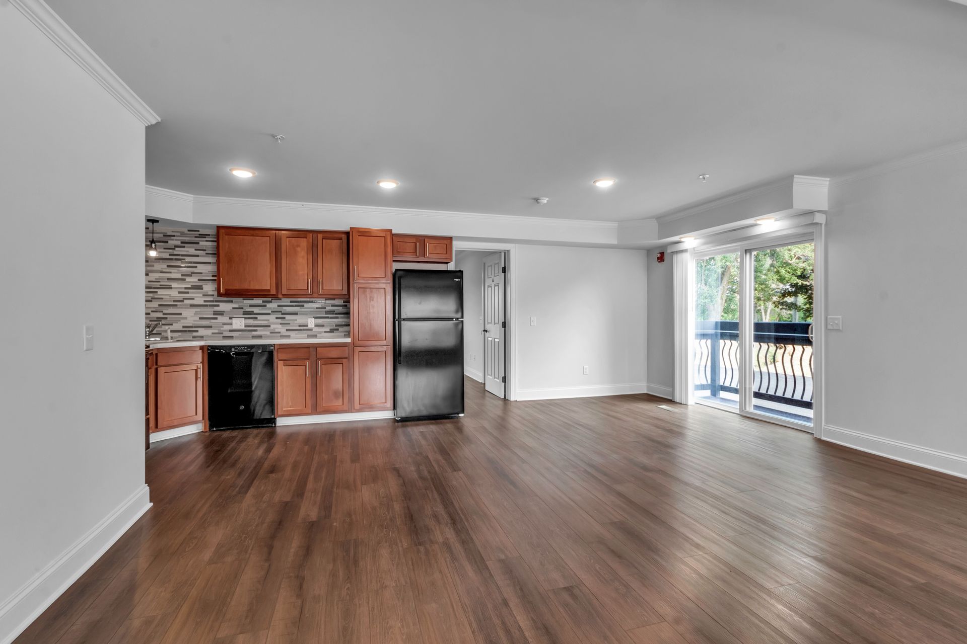 An empty apartment with hardwood floors , a refrigerator , and a sliding glass door.
