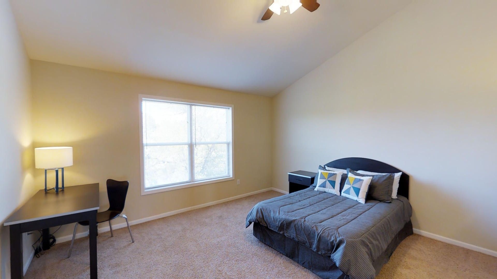 A bedroom with a bed , desk , lamp and window.