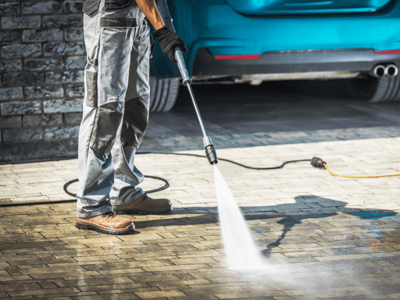 Person power washing a brick driveway near a teal car.