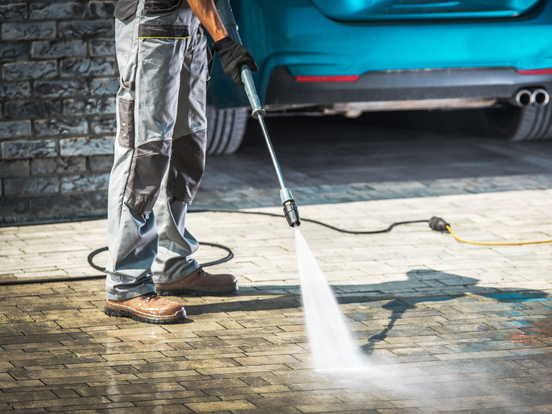 Person power washing a brick driveway near a teal car.