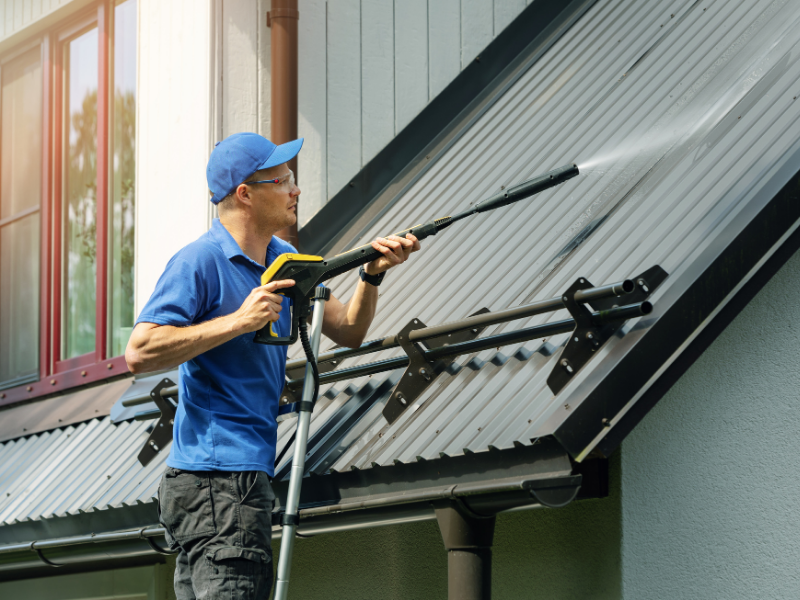 Man pressure washing a metal roof on a sunny day.