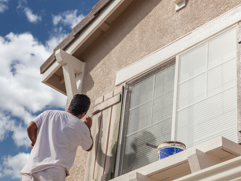 Man painting exterior trim of a house with a blue sky background.