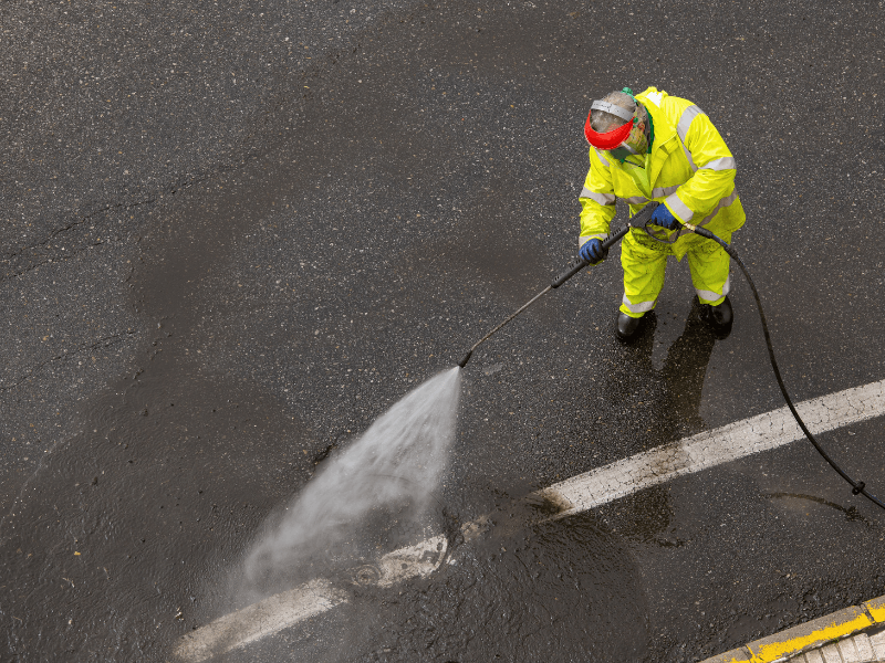 Person in yellow protective suit pressure-washing a dark parking lot surface.