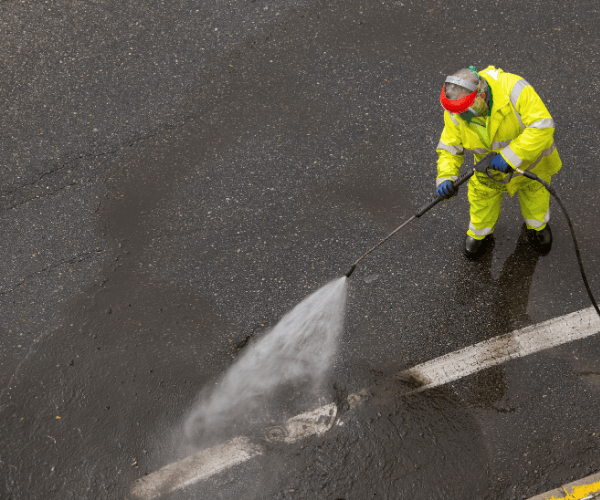 Person in yellow protective suit pressure-washing a dark parking lot surface.