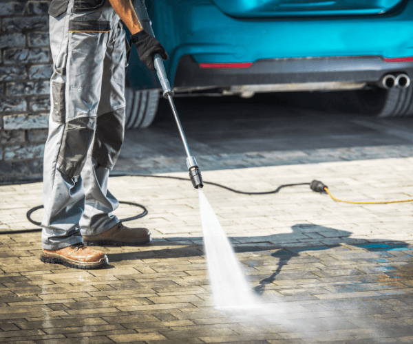 Person power washing a brick driveway near a teal car.
