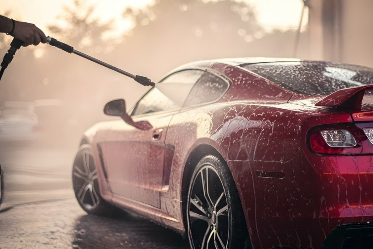 Red sports car being washed with a pressure washer; water and suds visible.