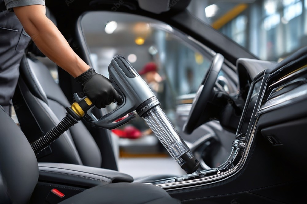 Person vacuuming the interior of a car. They wear gloves and hold a vacuum, cleaning the seats and console.