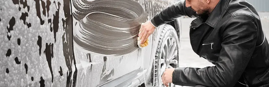 A man in a leather jacket is washing a car with a sponge.