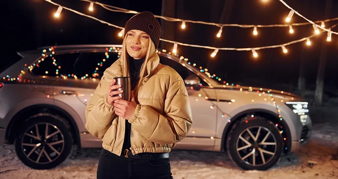 A woman is standing in front of a car holding a cup of coffee.