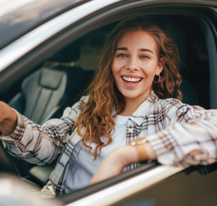Smiling woman sitting in a car, leaning out of the driver’s seat with a plaid shirt on.