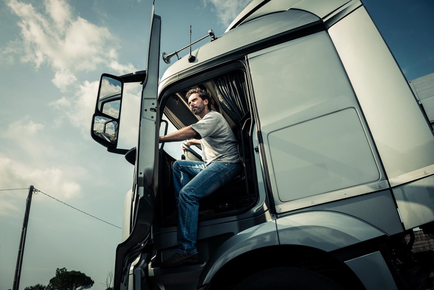 Man climbing into a white semi-truck cab, viewed from below against a cloudy sky