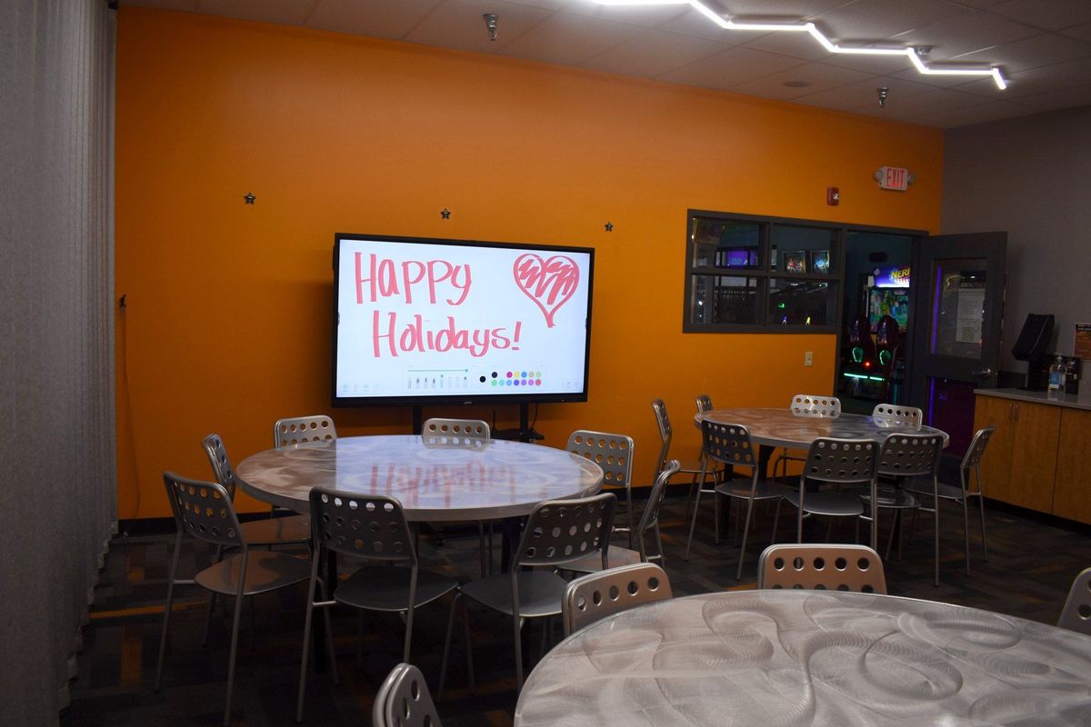 A party room at Zone 28 in Pittsburgh set with round tables, the smart board reading Happy Holidays, and the arcade through the window