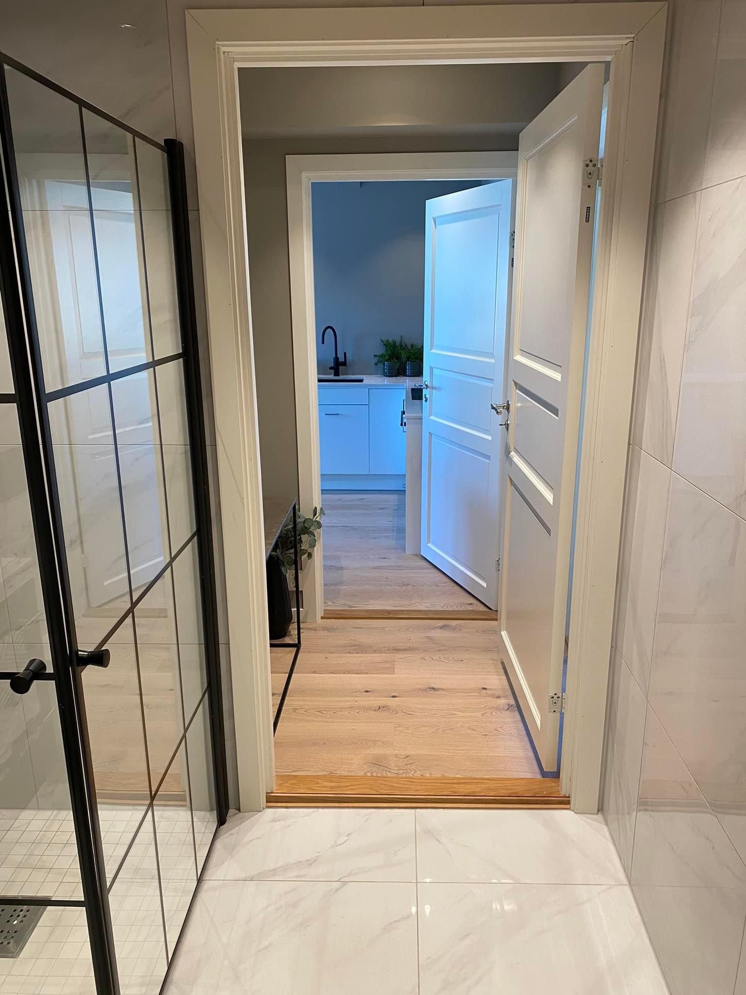 Baderomsgang med glassdusj, åpen hvit dør og flislagt gulv.